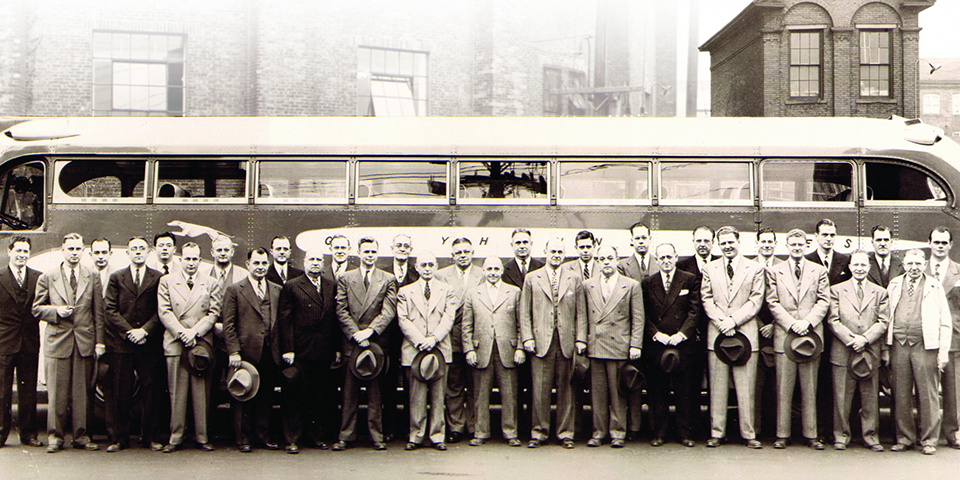 Archive photograph, a group of American manufacturers standing in front of a mid-century coach bus, suits and hats, black and white.