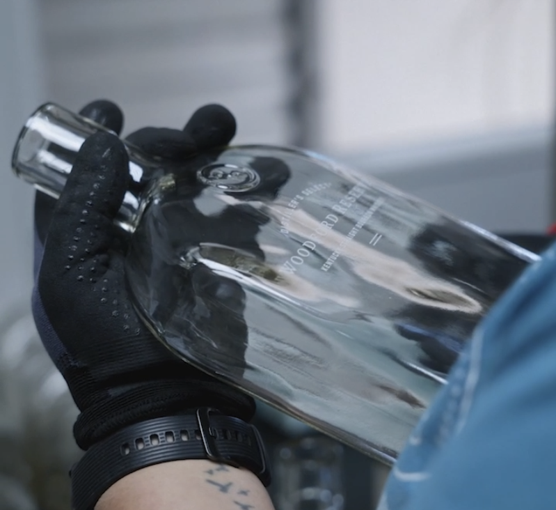 A gloved hand holds a Woodford Reserve glass bottle on the Chattanooga Labeling Systems decoration line, the embossed label finish visible on the bottle face.